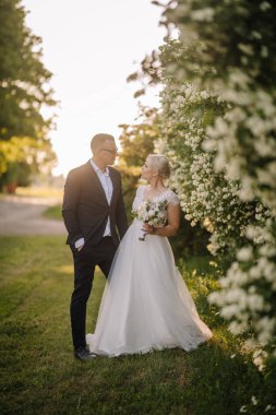 Beyaz elbiseli gelin, siyah takım elbiseli damadın yanında çiçek tutuyor, yemyeşil çiçeklerle çevrili, romantik bir düğün ortamı yaratıyor.