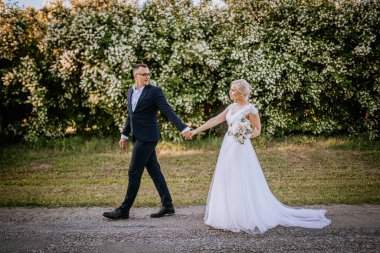 Düğün kıyafetli bir çift, canlı yeşillik ve çiçeklerle çevrili bir patikada birlikte yürüyor, romantik bir neşe ve bağ anını yakalıyorlar.