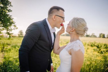 Romantik çift açık havada eğleniyor, canlı bir yeşillik ve açık bir gökyüzü arasında çilek paylaşıyor, aşkı ve mutluluğu simgeliyor.