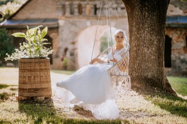 Zarif beyaz elbise giyen gelin, bir ağacın altında salıncakta sallanıyor. Arka planda yeşil ve kırsal bir bina var. Huzurlu bir düğün sahnesi oluşturuyor.