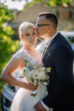 Zarif beyaz elbiseli gelin buketi tutarken, damat, canlı yeşilliklerle çevrili alnından öpüyor, doğada sevgi ve mutluluk anı sergiliyor.