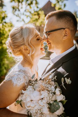 Çift doğal ortamda hassas bir anın tadını çıkarıyor. Gelin gülümsüyor ve elinde buket tutuyor. Düğün kutlamaları sırasında sevgi ve mutluluk sergiliyor.