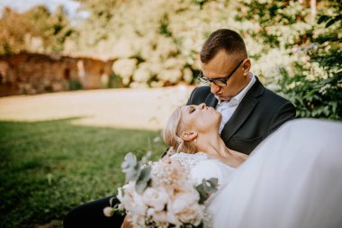 Zarif beyaz elbiseli gelin ve şık siyah takım elbiseli damat, açık havada, canlı yeşilliğin ortasında, güzel bir çevrede aşk ve romantizm gösteren hassas bir anı paylaşıyorlar.