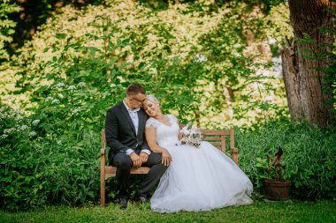 Yeşilliklerle çevrili, canlı bir bahçede bir bankta oturan yeni evli çift, doğada sevgi ve kutlamanın hassas bir anını yakalıyorlar.