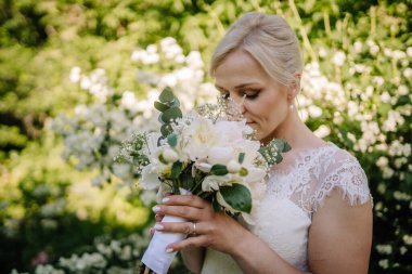 Zarif saç stiline sahip gelin, beyaz çiçekleri nazikçe tutuyor, canlı yeşilliğe daldırıyor, açık havada romantizm ve neşe uyandırıyor.