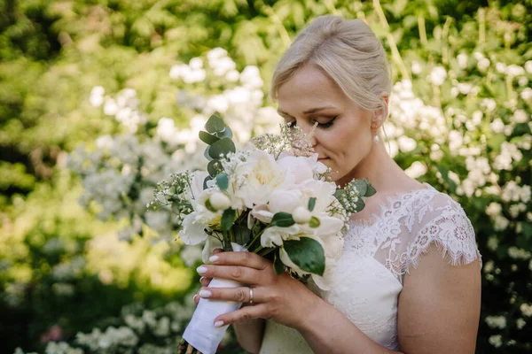 Zarif saç stiline sahip gelin, beyaz çiçekleri nazikçe tutuyor, canlı yeşilliğe daldırıyor, açık havada romantizm ve neşe uyandırıyor.