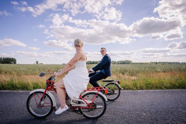 Düğün kıyafetleri içinde canlı yeşil tarlaları ve kabarık bulutları romantik ve kaygısız bir atmosfer yaratan neşeli bisiklet süren bir çift.