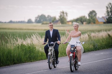 Mutlu çift manzaralı bir yolda bisiklet sürüyor, arka planda canlı yeşil alanlar ve ağaçlar, kaygısız ve romantik bir atmosferi somutlaştırıyor.