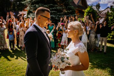 Düğün kıyafetli bir çift, mutlu misafirlerle çevrili, açık havada, neşeli bir anı paylaşıyor, canlı yeşillik ve sıcak güneş ışığı ile neşeli kutlamayı güçlendiriyor.