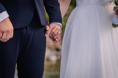 Resmi giyinmiş bir çiftin elleri sarmaş dolaş, sakin bir su gövdesinin yanında durur, dışarıda güzel bir ortamda aşkın ve birliğin özünü yakalar.