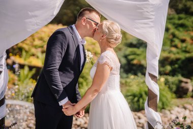 Düğün kıyafetleri içinde romantik bir çift güzel bir kemerin altında öpüşüyorlar, etrafları renkli çiçekler ve yeşilliklerle çevrili, sevgi ve neşe dolu