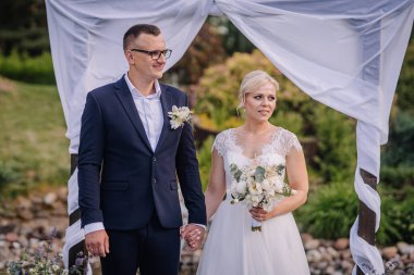 Çift güzel bir kemer altında bir arada durur, el ele tutuşur ve gülümser, canlı çiçekler ve yeşilliklerle çevrili, aşk ve kutlama anlarını yakalar.