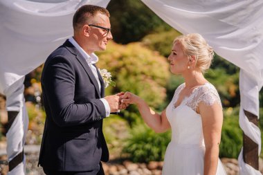 Düğün kıyafetli bir çift, güzel yeşillik ve dekoratif kumaşlarla açık havada evlilik yemini ederken özel günleri için romantik bir ortam yaratıyor.