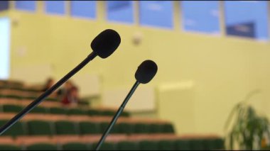 Konferans salonunda iki mikrofon belirgin bir şekilde gösteriliyor, boş koltuklar ve yumuşak ışıklandırma odaklı bir atmosfer yaratıyor, kamera detaylar için pürüzsüz bir şekilde yakınlaşıyor