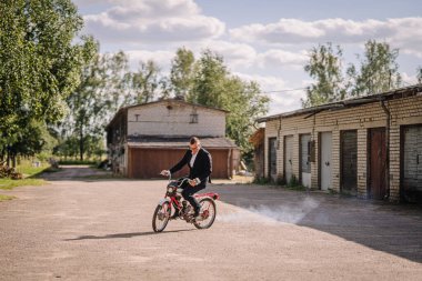 Erkek motosikletçi, ağaçlar ve binalarla çevrili taşra bölgesinde çakıl yolda ustaca motosiklet sürüyor ve açık hava macerasının özünü yakalıyor.