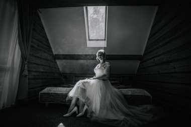 Beyaz elbiseli zarif gelin, kırsal bir odada yatakta oturuyor. Tavan penceresinden gelen yumuşak doğal ışıkla aydınlanıyor. Huzurlu ve romantik bir ortam yaratıyor.