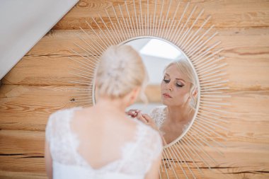 Gelin, şık, yuvarlak bir aynanın önünde makyaj yapmaya odaklanmış, ahşap duvarlar sıcak bir atmosfer yaratıyor, düğün hazırlıklarını gözler önüne seriyor.