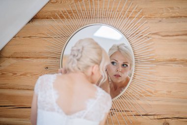 Beyaz elbiseli zarif gelin, güneş ışığı desenli yuvarlak aynaya bakar, kırsal ahşap duvarlara yaslanır, sakin ve samimi bir atmosfer yaratır.