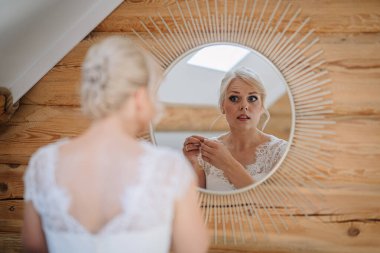 Gelin düğün günü için hazırlanıyor, yuvarlak aynaya bakarken küpeleri ayarlıyor, kırsal ahşap duvarlar sıcak bir atmosfer yaratıyor.