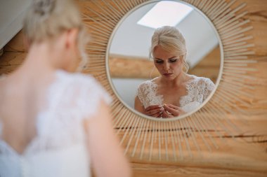 Sarışın gelin dantelli gelinlikle, yuvarlak bir aynada mücevherleri ayarlarken görülüyor, kırsal ahşap duvarlar sıcak bir atmosfer yaratıyor, zarafet ve hazırlık anlarını yansıtıyor.