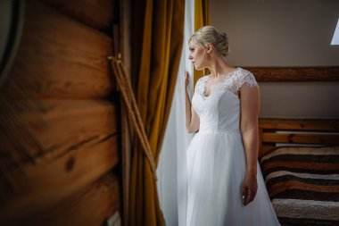 Güzel beyaz gelinlikli gelin pencerenin yanında duruyor, yumuşak ışıkla yıkanmış, ahşap duvarlar ve sıcak perdelerle huzurlu ve samimi bir atmosfer yaratıyor.