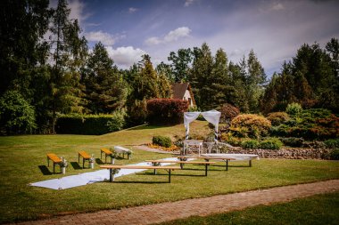 Güzel bir açık hava düğün sahnesi, beyaz bir kemer, ahşap banklar, yeşil bir çimenlikte masalar, renkli ağaçlar ve çalılarla çevrili, romantik bir kutlama için mükemmel.