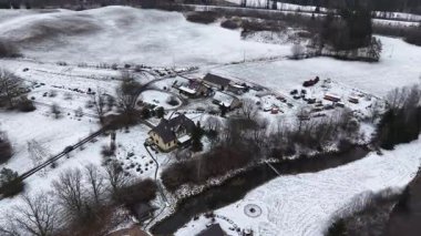 Cesis, Letonya - 21 Kasım 2025: Hava görüntüsü, karla kaplı tarlalarla çevrili bir nehir kıyısında, manzarayı düzgünce gösteren bir çiftlik evi yakalıyor.