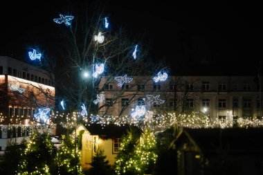 Valmiera, Letonya - 01 Aralık 2025: Şenlikli pazar sahnesinde Noel ışıkları, parıldayan süslemeler ve ağaçlarla çevrili büyüleyici bir ahşap kulübe yer alıyor