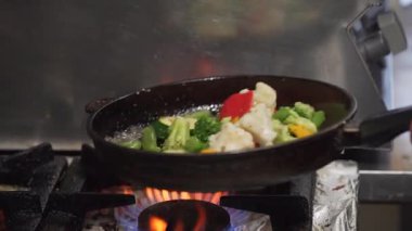 Dinamik aşçılık sahnesinde rengarenk sebzeler alevlerin üzerine fırlatılıyor. Mutfak hareketlerini ve canlı renkleri vurguluyorlar.