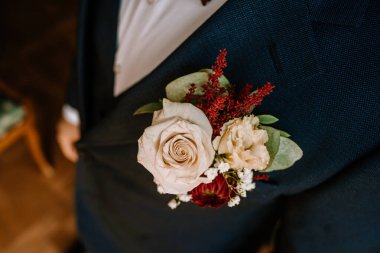 Krem rengi gül ve canlı kırmızı aksanlı çiçekli yaka çiçekleri. Özel dikim bir takım elbiseyle süslenmiş. Kutlamalar için resmi kıyafetlere zarafet ve cazibe katıyor.