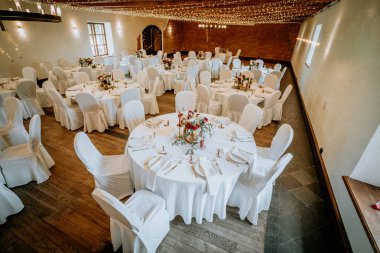 Şölen salonunda beyaz çarşaf, çiçek aranjmanı ve çevre aydınlatması giyinmiş yuvarlak masalar yer alıyor ve özel etkinlikler ve kutlamalar için hoş bir ortam yaratıyor.
