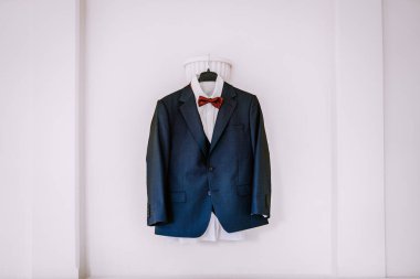 Papyonlu resmi takım elbise, el değmemiş beyaz bir duvara asılarak zarif bir şekilde sergileniyor, sofistike moda ve kutlamaya hazır olma durumu vurgulanıyor.