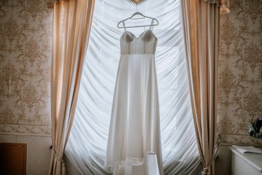 Beyaz gelinlik pencereden zarif bir şekilde sergileniyor, yumuşak ışıkla yıkanmış, sahneyi çerçeveleyen hassas perdeler, gelin anları için romantik bir ortam yaratıyor.