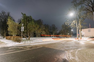 Rujiena, Letonya - 27 Kasım 2025: Şehir gece sahnesinde karlı bir sokakta aydınlatılmış ağaçlar ve ışık yolları tasarım için geniş bir kopya alanına sahip.