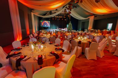 Kutlama için şölen salonu seti, beyaz çarşaflarla süslenmiş yuvarlak masalar, yumuşak ışıklandırma, ve bir Noel ağacı, samimi ve davetkar bir ortam yaratıyor.