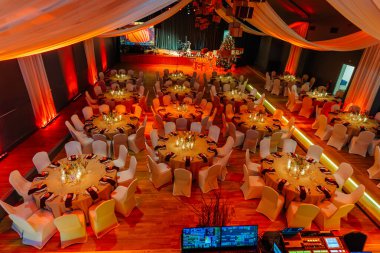 Şölen salonunda beyaz masa örtüleri ve zarif süslemeleri olan yuvarlak masalar, şenlikli bir ortam için sıcak aydınlatılmış, düğünler ve kutlamalar için ideal.