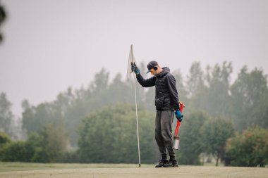 Siyah ceket ve eldiven giyen personel, golf sahasındaki bayrak direğini yeşil renge göre ayarlamaya, arka planda ağaçlar bulunarak pistin korunması yönündeki kararlılığı vurgulamaya odaklanıyor.