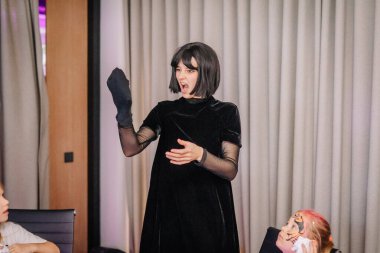 Riga, Letonya - 24 Ekim 2025 Cadılar Bayramı partisi. Siyah elbiseli kadın sanatçı, modern kapalı ortamlarda seyircileri eğlendirmek için kukla kullanır.