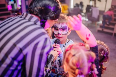 Riga, Letonya - 24 Ekim 2025 Cadılar Bayramı partisi. Yüzü boyalı çocuk canlı çocukların partisinde renkli süslemelerle mikrofona şarkı söylüyor.