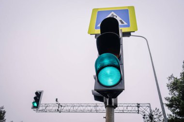 Bulutlu bir gökyüzüne karşı aydınlatılmış yeşil trafik ışığı, araçların ilerleyeceğini gösteriyor, yaya geçidi işareti ile, kentsel güvenlik ve ulaşım dinamiklerini vurguluyor