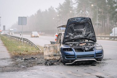 Yol kenarında terk edilmiş, kaputu açık, yoldan geçen araçlarla çevrili, araç yangınlarının ve yol güvenliği endişelerinin sonuçlarını gözler önüne seriyor.
