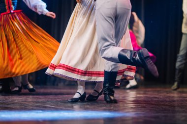 Renkli giyinmiş Letonyalı halk dansçıları sahnede sahne alarak kültürel geleneklerini ve ritmik hareketlerini enerjik bir ortamda vurguluyorlar.