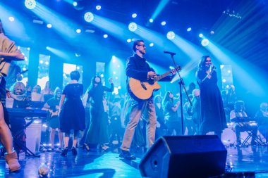 Valmiera, Letonya - 29 Aralık 2025: Yamaha. Erkek gitarist, kadın şarkıcı ve koroyla renkli ışıklarla dolu canlı bir atmosferde sahneye çıkıyor.