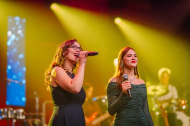 Valmiera, Letonya - 29 Aralık 2025: Yamaha. Kadın vokalistler, renkli ışıklar ve enerjik bir atmosfer yaratan canlı bir grupla sahnede tutkuyla şarkı söylüyorlar.