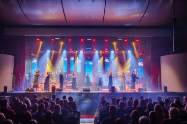 Valmiera, Letonya - 29 Aralık 2025: Yamaha. Sahnede canlı ışıklar eşliğinde canlı performans sergileyen çeşitli şarkıcılar ve konferans salonunda meşgul bir seyirci grubu.