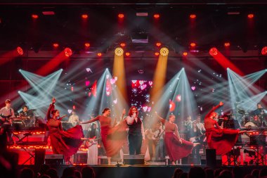 Valmiera, Letonya - 29 Aralık 2025: Yamaha. Sahnede kırmızı elbiseli ve renkli ışıklarla dans eden dansçılar enerjik bir ortam yaratıyorlar.