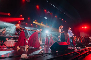 Valmiera, Letonya - 29 Aralık 2025: Yamaha. Kırmızı elbiseli dansçılar sahnede canlı ışık ve enerjik bir atmosfer yaratan müzisyenlerle performans sergiliyorlar.