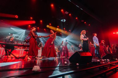 Valmiera, Letonya - 29 Aralık 2025: Yamaha. Zarif kırmızı elbiseler giyen sanatçılar sahne arkasında dramatik ışıklandırma altında müzisyenlerle dans ediyorlar.