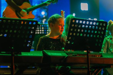 Valmiera, Letonya - 29 Aralık 2025: Yamaha. Kız renkli sahne ışıkları ve müzisyenlerle canlı müzik performansı sırasında klavye çalmaya odaklanmış.