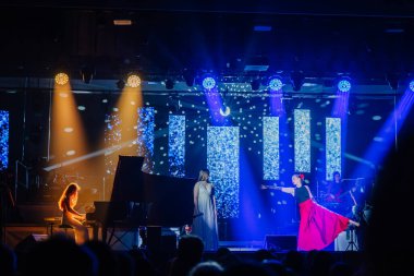 Valmiera, Letonya - 29 Aralık 2025: Yamaha. Kırmızı elbiseli şarkıcı, renkli sahne ışıkları ve dinamik görsellerle aydınlatılmış piyanist çalarken seyirciyi büyülüyor.